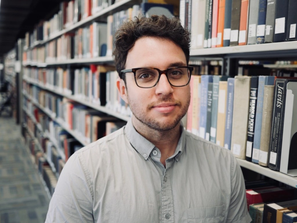 Headshot of Dave Rodriguez. He has brown hair, glasses, and stands in front of a shelf of books.
